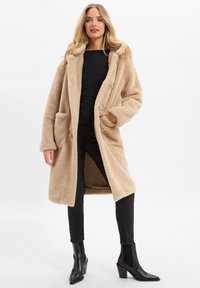 Frau in einem beigen langen Teddy-Mantel, schwarzem Ripp-Top, schwarzen Skinny-Jeans und schwarzen Absatz-Stiefeletten, die vor einem weißen Hintergrund steht.