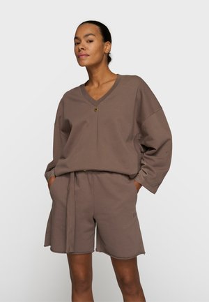 Kobieta ubrana w luźną brązową bluzkę z dekoltem w serek i długimi rękawami oraz pasujące krótkie spodenki do kolan, stojąca z rękami w kieszeniach na jednolitym tle.