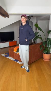 Joven con sudadera azul marino con capucha, pantalones de chándal grises y gorra, de pie con las manos en los bolsillos en una sala de estar moderna con sofá y una maceta grande con planta.
