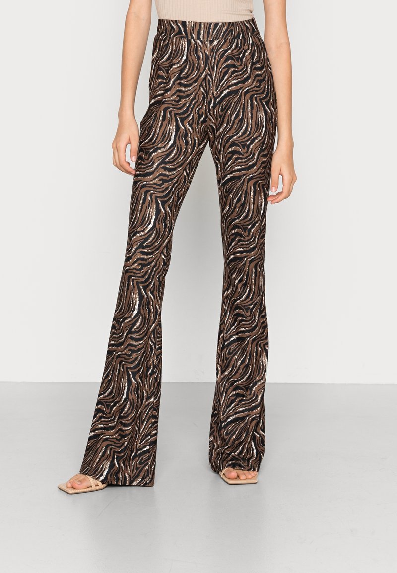 Pantalon évasé taille haute à imprimé zébré marron et noir, porté avec un haut couleur nude et des sandales beige à bout carré, sur un modèle debout devant un fond uni.