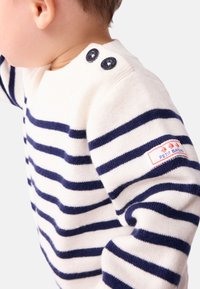 Maglione bianco con righe orizzontali blu navy, caratterizzato da un collo rotondo, due bottoni blu navy sulla spalla e una piccola etichetta logo rossa sulla manica.