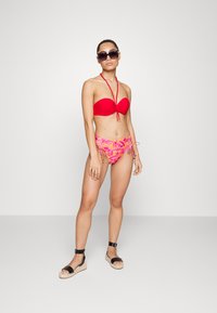 Top de biquíni bandeau vermelho com laços de halter e parte de baixo de biquíni floral rosa. A modelo usa óculos escuros escuros e sandálias espadrilles pretas.