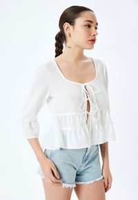 Blusa blanca, ligera, con escote cuadrado, cierre con cordón y dobladillo con volantes. Combinada con pantalones cortos de mezclilla claros.