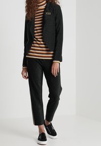 Blazer noir étincelant avec col cranté, associé à un col roulé rayé multicolore et un pantalon noir fuselé avec des bandes latérales.