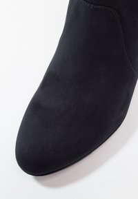 Gros plan sur l'avant d'une botte cheville en daim noir avec des coutures visibles sur fond blanc.