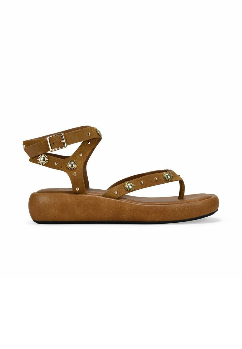 Sandalo in suede marrone con cinturino alla caviglia, caratterizzato da borchie decorative in metallo e dettagli floreali, su una suola piatta e sagomata.