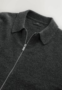 Pulover gri din fleece cu fermoar, guler ascuțit, fermoar argintiu și branding minim. Textură moale cu un finisaj neted.