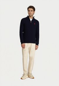 Mladý muž v námořnickém modrém svetr s pleteným vzorem a polozipem, béžových kalhotách a béžových teniskách, stojící na pozadí čistě bílé stěny.