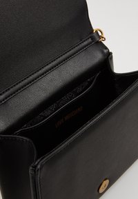 Czarna torebka skórzana Moschino otwarta, ukazująca wewnętrzną kieszeń oraz metalowy zamek magnetyczny w złotym odcieniu na przedniej klapie.