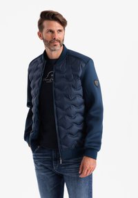 Veste d'aviateur marine avec détails matelassés, poignets côtelés et fermeture éclair. Portée par-dessus un t-shirt noir, associée à un jean en denim.
