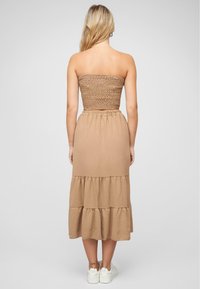 Bruine gelaagde maxi-jurk met een gesmokte buste, voorzien van een relaxed fit en elastische tailleband, gecombineerd met witte sneakers.