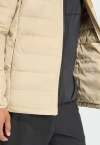 Beige padded jacket open to show black inner lining and printed care labels, worn with black pants by a person’s left hand.