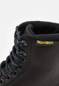 Svart lær ankle boot med lodden svart foret, seks svarte hull, mørk snøring og "WarmWair"-merke i gult sydd på siden.