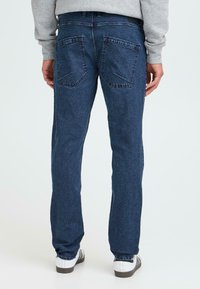 Blugi din denim albastru închis cu un croi drept, având două buzunare pe spate și o textură textilă subtilă. Purtați cu adidași albi.