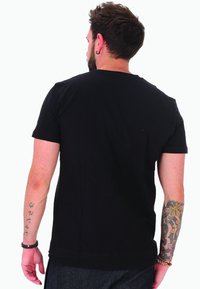 Uomo con i capelli corti e scuri e tatuaggi su entrambe le braccia, che indossa una maglietta nera semplice, di spalle su uno sfondo bianco.