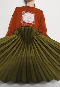 Sudadera de color óxido con diseño gráfico; falda plisada en verde oliva con tela texturizada y cinturilla fruncida, creando un contraste de estilos.