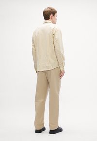 Lichtbeige geblokt overhemd met lange mouwen en een spread collar, gecombineerd met lichtbeige broek en zwarte schoenen. Effen achtergrond.