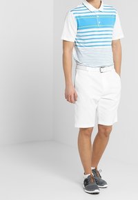 Homme portant un short blanc, une ceinture blanche, un polo à rayures bleues et blanches, et des chaussures de sport grises, debout dans un espace neutre.