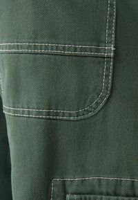 Tmavě zelená tkanina s texturovaným tkaním, se kontrastním bílým prošíváním a zakřivenou kapsou na pravé straně.