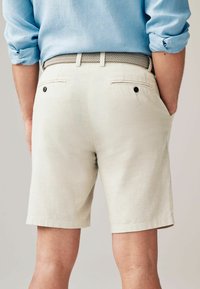 Pantaloni scurți din in bej, cu două buzunare la spate, unul dintre ele având un nasture. Țesătura are o textură netedă, tăiată la lungimea genunchiului.