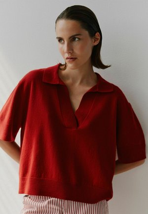 Suéter corto de manga corta rojo con cuello en V y un collar, que presenta un tejido texturizado. Combinado con pantalones a rayas.