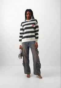 Sweatshirt rayé noir et blanc avec un petit détail de rose rouge, associé à un pantalon cargo décoloré avec plusieurs poches. Modèle tenant un sac gris.