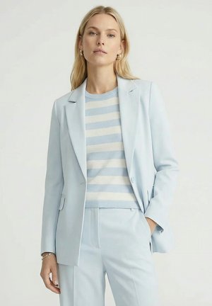 Frau, die einen hellblauen Anzug mit einem gestreiften beigefarbenen und blauen Pullover trägt, steht mit einer Hand in der Tasche vor einem schlichten Hintergrund.