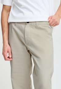 Beige bukser med en jevn tekstur, som har beltehemper, frontglidelås og sidelommer, båret under en hvit skjorte.