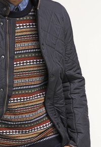 Veste matelassée bleu marine avec des accessoires en bronze, portée sur un pull multicolore à motifs, comprenant des rayures horizontales et des motifs géométriques.