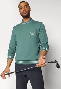 Pullover verde a maniche lunghe con colletto, dotato di una tasca sul petto e dettagli a righe. Il modello tiene un bastone da golf.