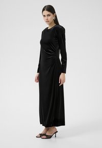 Robe longue en velours noir à manches longues avec un détail de fronces sur le côté, coupe ajustée, col haut et fente latérale pour faciliter les mouvements, associée à des talons noirs.