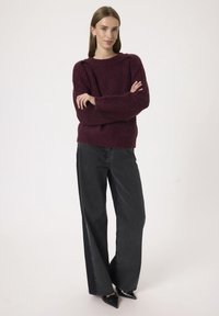 Maglione bordeaux a coste con un ampio scollo e maniche lunghe, abbinato a jeans neri a gamba larga e tacchi a punta neri. Sfondo neutro.