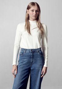Weißes Langarmoberteil, kombiniert mit hochgeschnittenen blauen Jeans aus Denim. Das Oberteil hat eine taillierte Passform; die Jeans haben einen entspannten Schnitt und sichtbare Nähte.