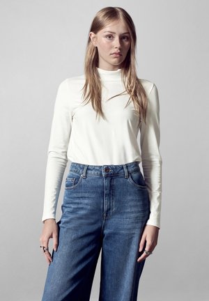 Witte langemouwen top in combinatie met hoge taille blauwe spijkerbroek. De top heeft een getailleerd ontwerp; de jeans hebben een ontspannen pasvorm en zichtbare stiksels.
