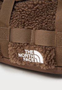 Brown fleece fabric with a textured surface, featuring a white logo "THE NORTH FACE" and brown webbing straps for secure fastening.