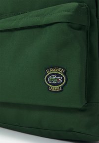 Sac à dos en toile verte avec une surface texturée et un logo Lacoste brodé bien en vue, représentant un crocodile et le texte "PARIS".