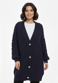 Granatowy kardigan dziany z głębokim dekoltem w serek, grubymi rękawami w warkoczowy splot i sześcioma dużymi ozdobnymi guzikami z przodu.
