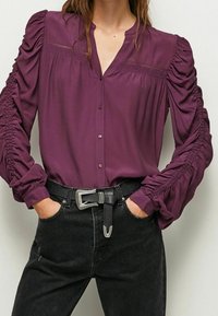 Blusa viola con bottoni davanti, maniche arricciate e dettagli di cucitura decorativa. Abbinata a jeans neri e una cintura testurizzata.