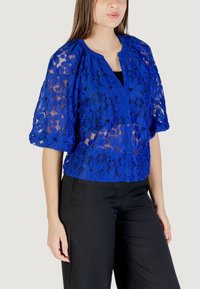Blusa in pizzo blu reale con motivi floreali, maniche corte a palloncino, scollatura a V e dettagli con bottoni, abbinata a pantaloni neri.