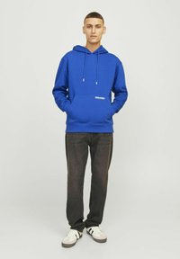 Felpa con cappuccio blu, dotata di tasca frontale, lacci e logo bianco. Abbinata a jeans scuri e sneaker bianche con dettagli neri.