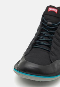 Black sneaker with a smooth leather toe cap, textured fabric upper, and blue accent on the sole; features black laces and a Camper logo.