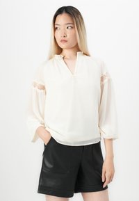 Blusa color crema con colletto alla mandarina, dettagli in pizzo sulle maniche e tessuto morbido e leggero. Abbinata a pantaloni corti neri in pelle.