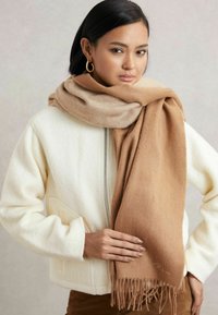 Fular din lână maro cu margini festonate, lăsat lejer, asortat cu o geacă albă texturată. Textura netedă și tonurile calde domină designul.