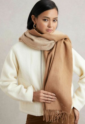 Brun ullscarf med fransar, hängande löst, ihop med en vit texturerad jacka. Slät textur och varma nyanser dominerar designen.