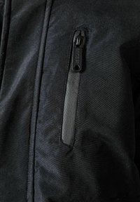 Vêtement d'extérieur en tissu noir avec une poche poitrine zippée. Texture lisse, fermeture éclair robuste et détails de couture à l'avant.