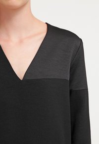 Top noir avec un décolleté en V, ayant une texture mate dans la partie supérieure et une finition lisse dans la partie inférieure. Les bords des manches sont simples.