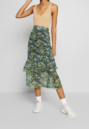 Personne portant un haut sans manches en tricot beige, une jupe midi à imprimé floral superposée avec des fleurs vertes et bleues, des chaussettes blanches et des baskets blanches, se tenant debout.