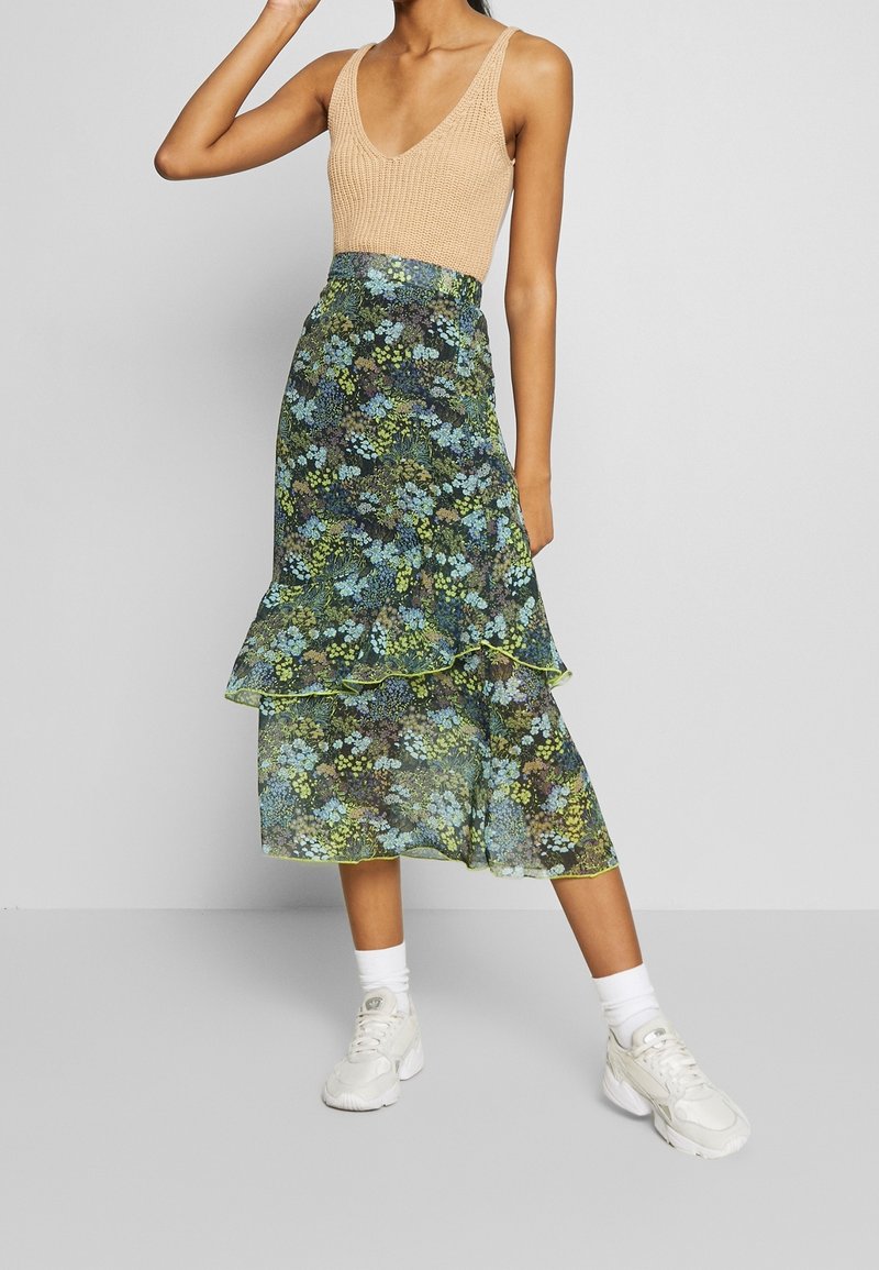 Personne portant un haut sans manches en tricot beige, une jupe midi à imprimé floral superposée avec des fleurs vertes et bleues, des chaussettes blanches et des baskets blanches, se tenant debout.
