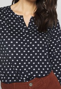 Navybluse mit weißen ovalen Punkten, ausgestattet mit einem V-Ausschnitt und gerafften Details. Kombiniert mit rostfarbenen Cordhosen.