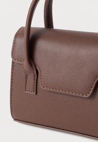 Brun läderhandväska med ett kort handtag och scalloped lock, detaljerad sömnad på kanterna, framför en enkel ljus bakgrund.